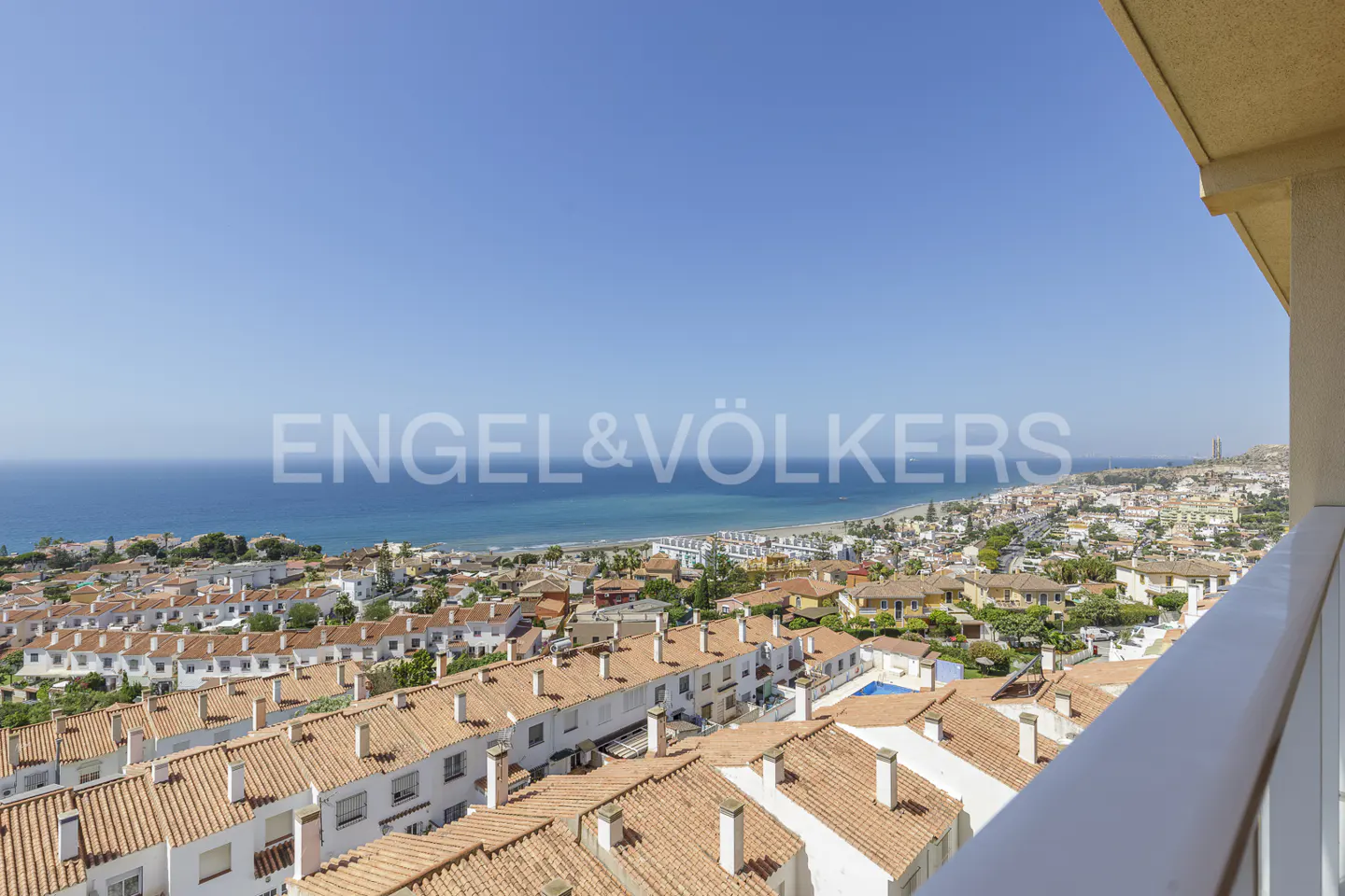 Elevated view from a balcony overlooking a coastal town with white houses, terracotta roofs, and the blue Mediterranean Sea.