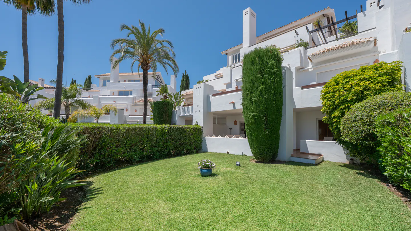 Lush green lawn in front of white Mediterranean-style homes with palm trees under a clear blue sky.