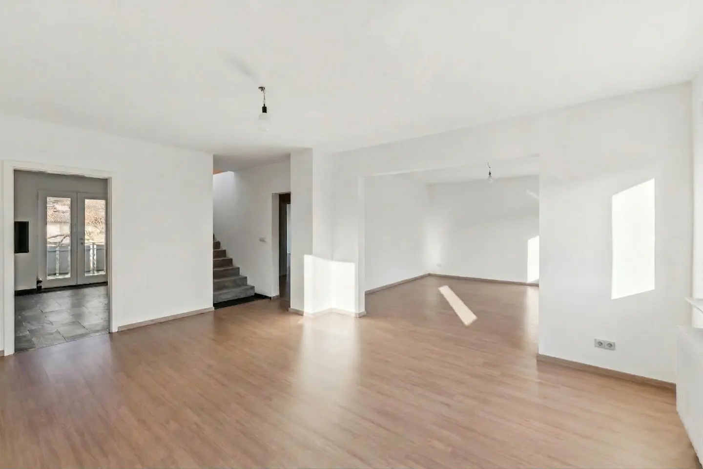 Bright, empty room with wood floors and white walls. A staircase and doorway to a patio are visible.