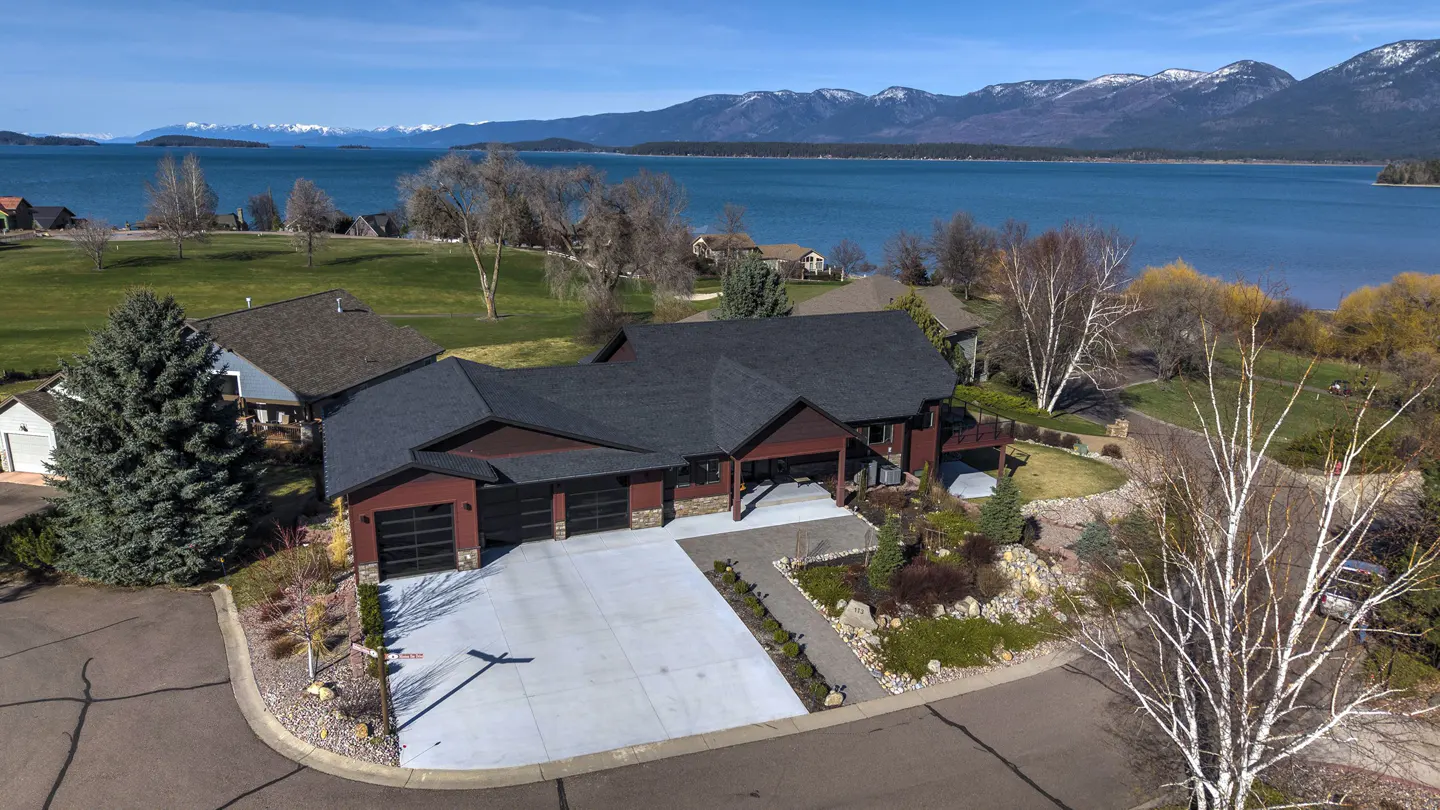 Aerial view of a modern, red house with a black roof and three-car garage, set against a backdrop of a blue lake and snow-capped mountains.