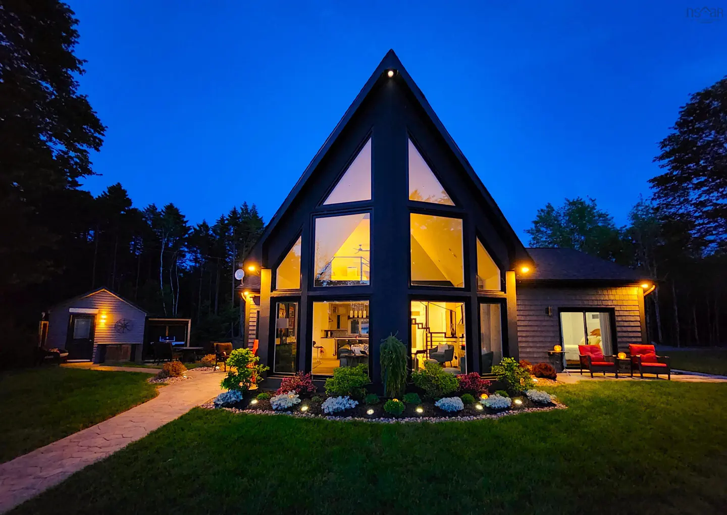 Exterior view of a modern A-frame house at dusk, with large windows, a stone path, and manicured landscaping.