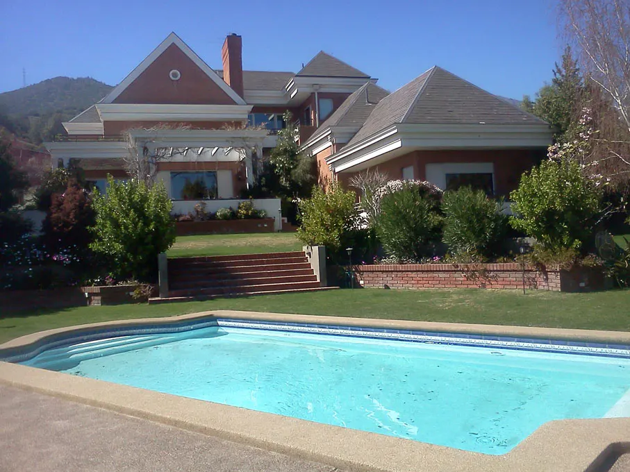 A large brick house with a pool in the foreground. The pool is blue and surrounded by a concrete deck. There are green trees and grass around the house.
