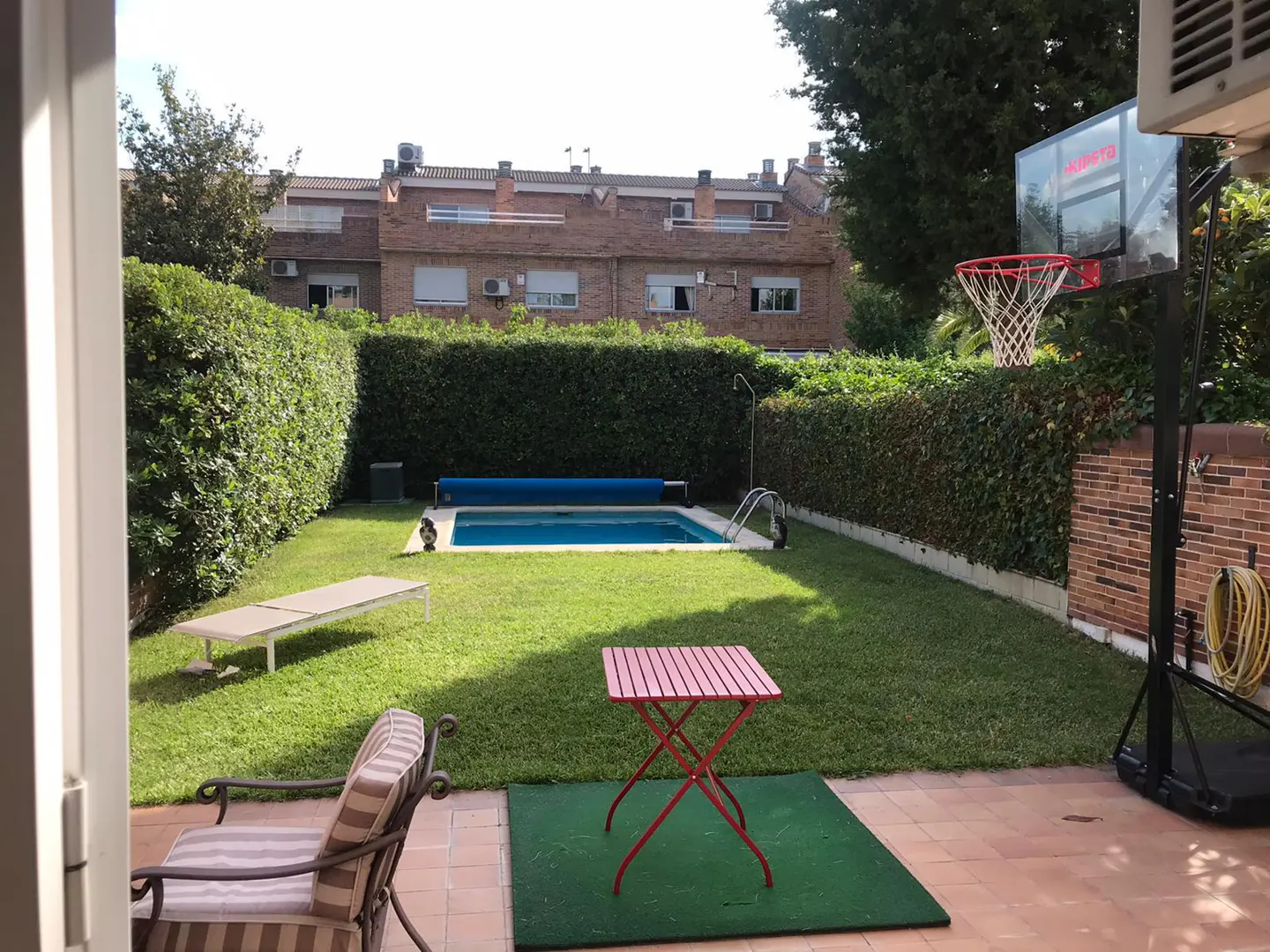 A backyard with a pool, green grass, and a red folding table on a green mat. A basketball hoop stands on a brick patio. Hedges line the yard.