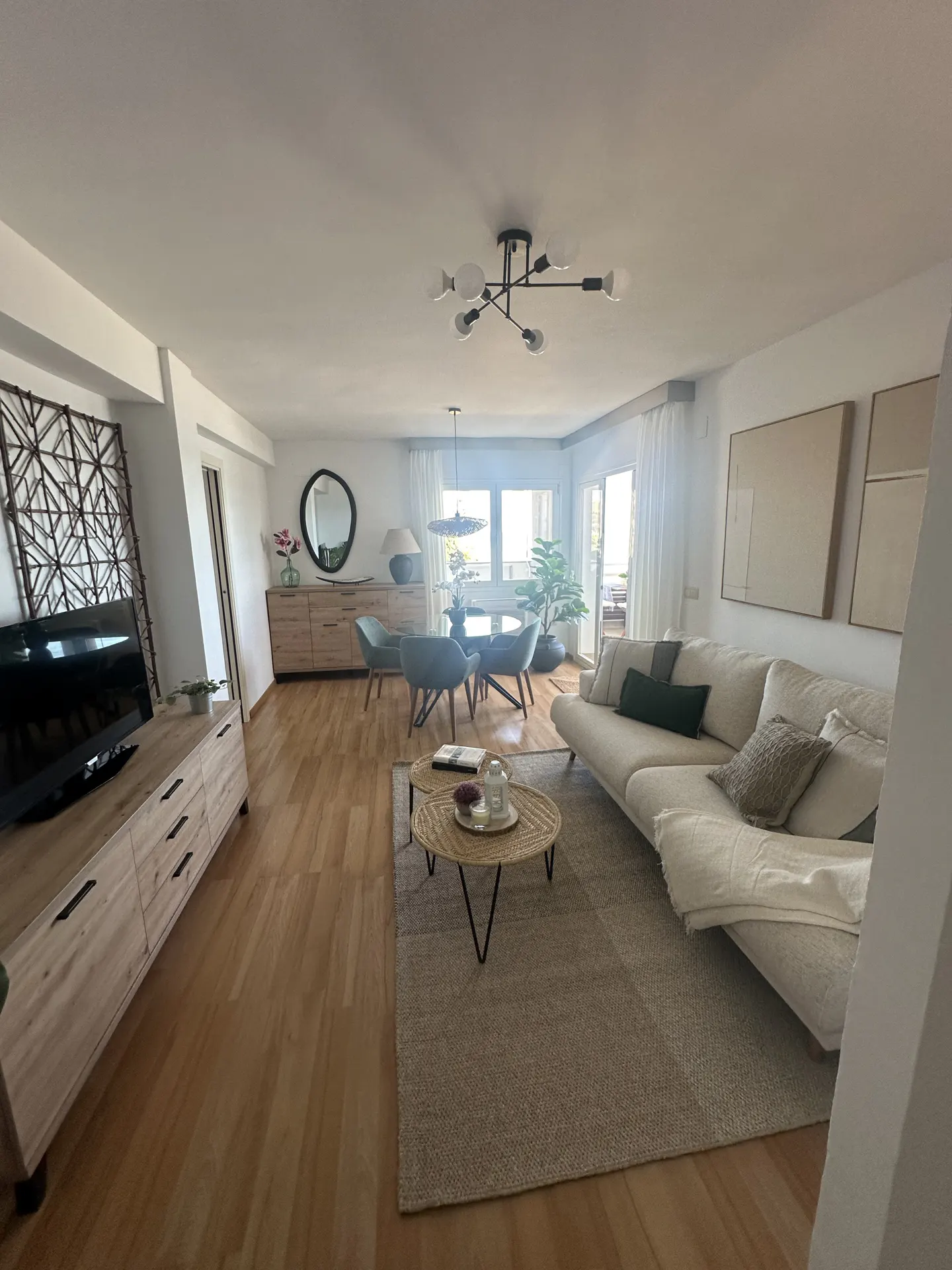 A bright living room with a beige sofa, round dining table, and light wood floors. A TV sits on a wooden cabinet.