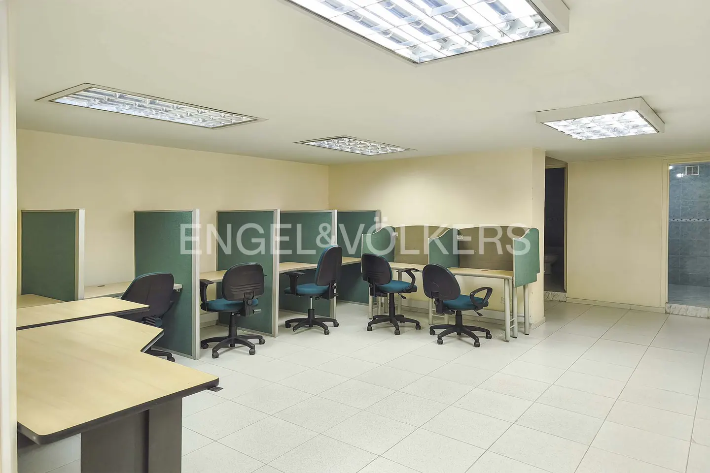 Office space with desks, teal chairs, and green partitions under fluorescent lights.