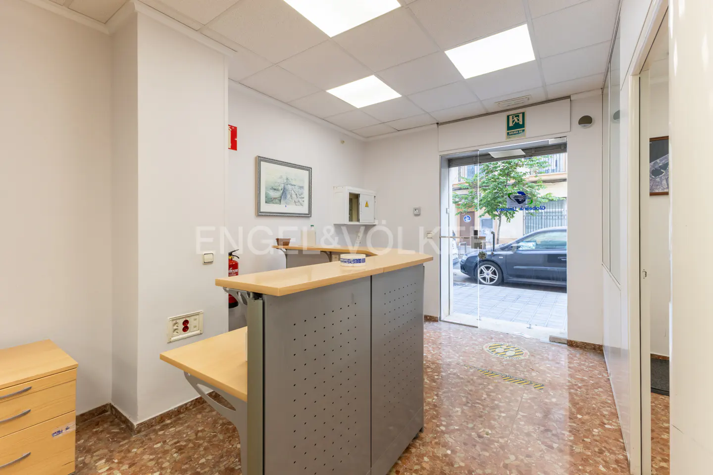 Office interior with a gray reception desk, light wood countertop, and a view of the street through the glass door.