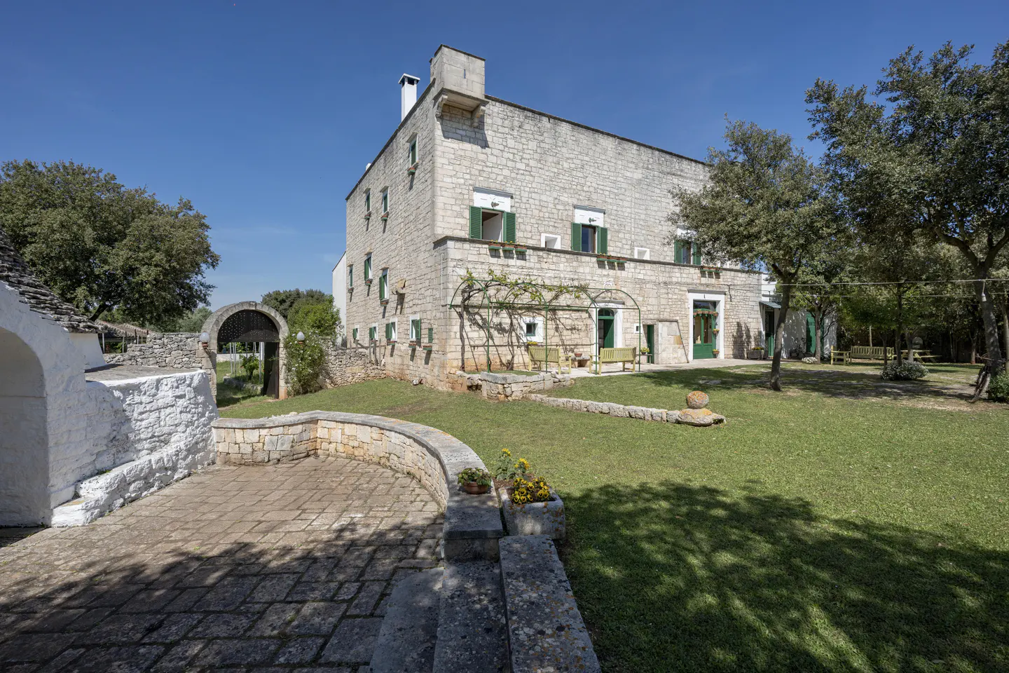 Exterior of a stone house with green shutters, a patio, and a large lawn under a blue sky.