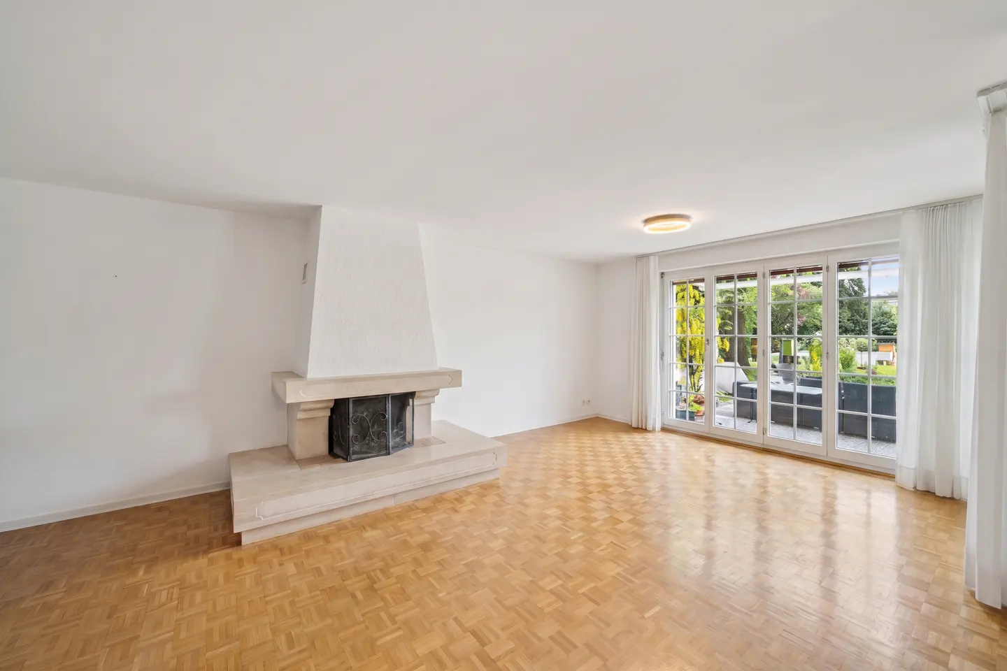 Bright, empty living room with parquet floors, white walls, and a stone fireplace. Large glass doors lead to a patio and garden.