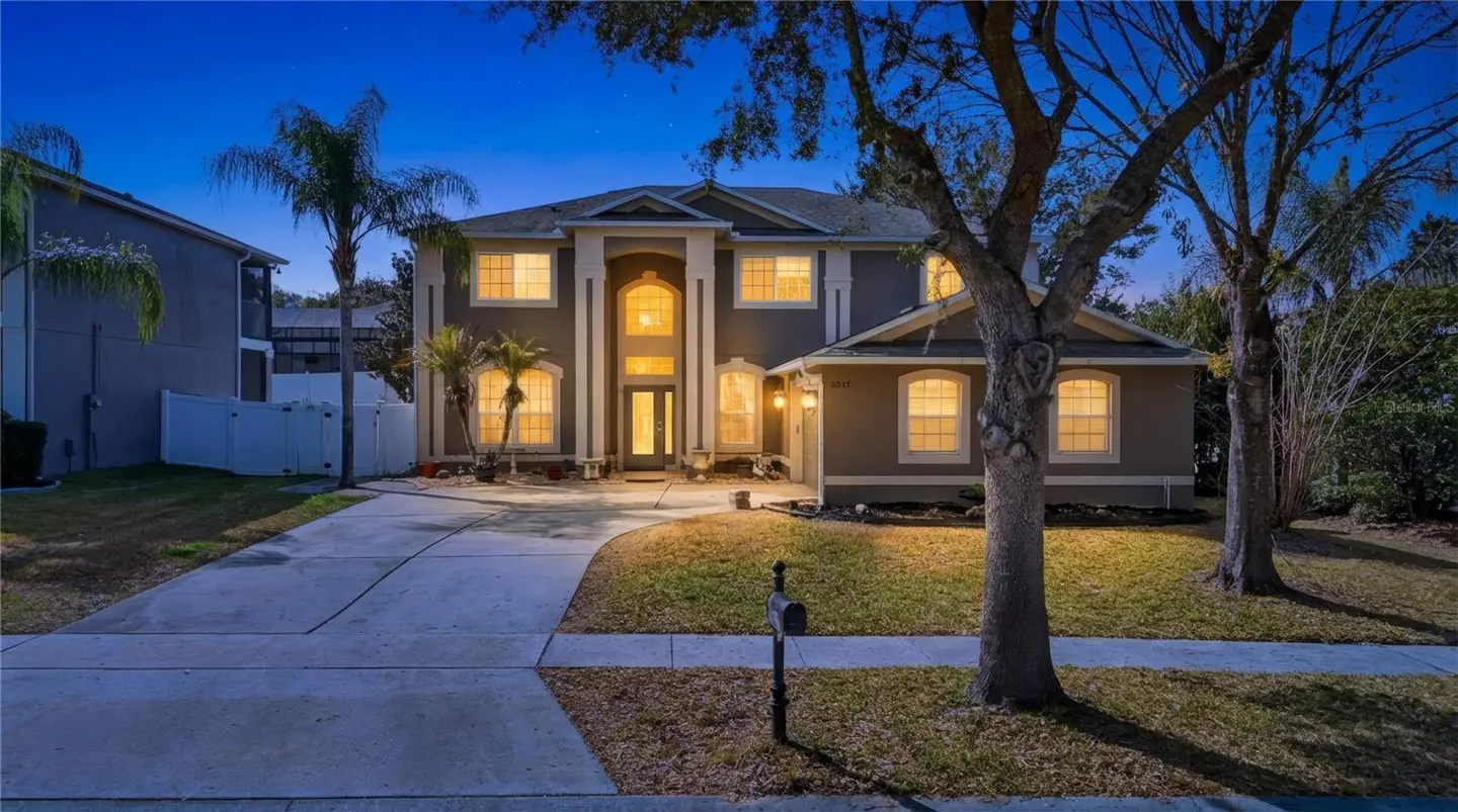 Two-story gray house with white trim, illuminated windows, and a long driveway at dusk. Palm trees and a mailbox are in the front yard.