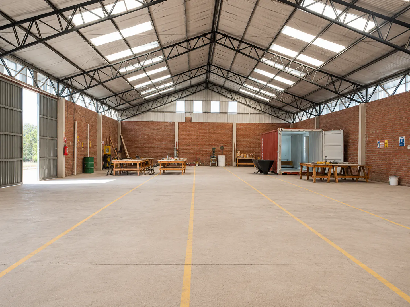 Spacious industrial warehouse with brick walls, concrete floor with yellow lines, skylights, and wooden workbenches.