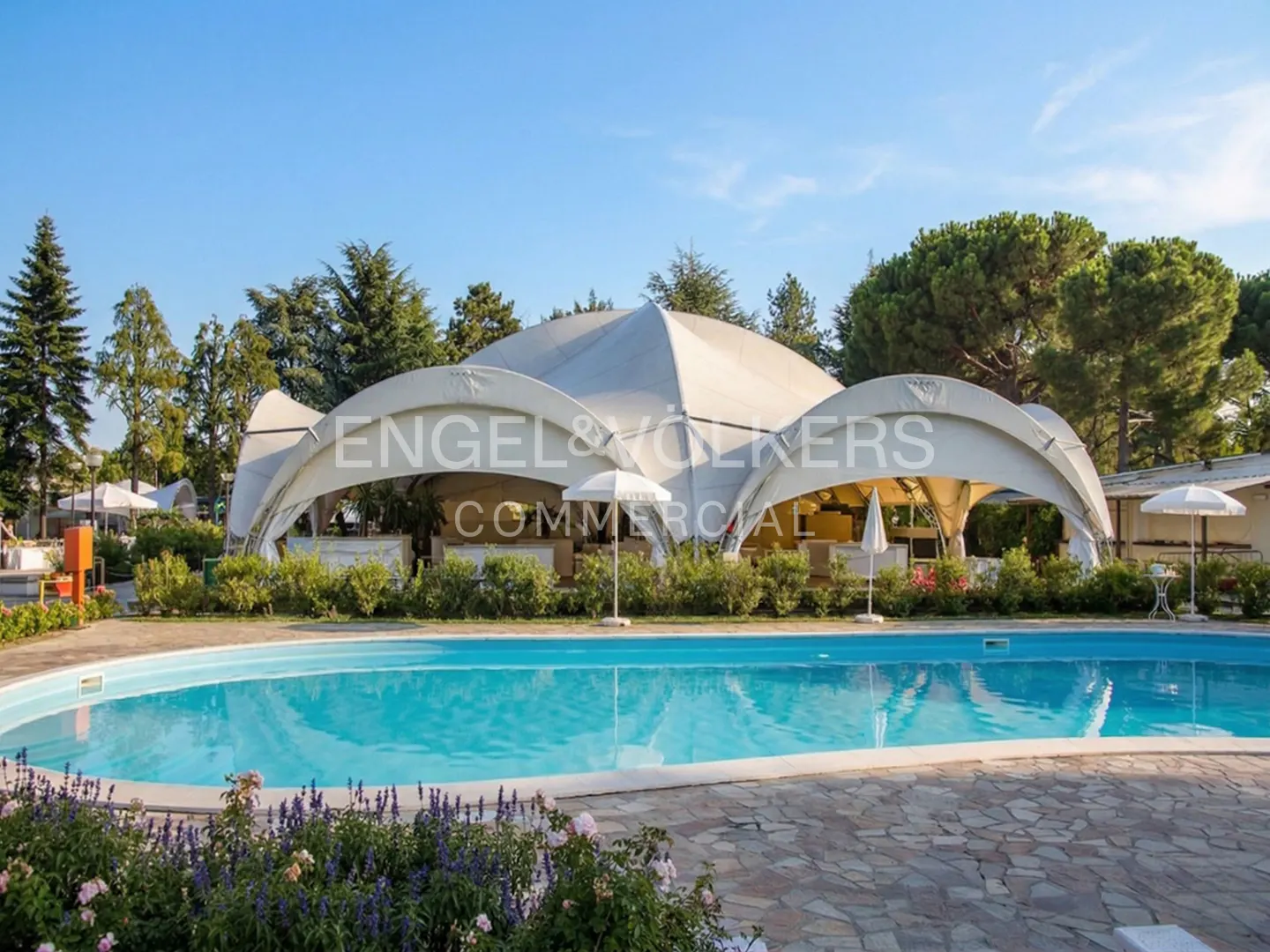 Outdoor pool with a white event tent, tables, and chairs. Flowers line the pool's edge. Trees and blue sky in the background.