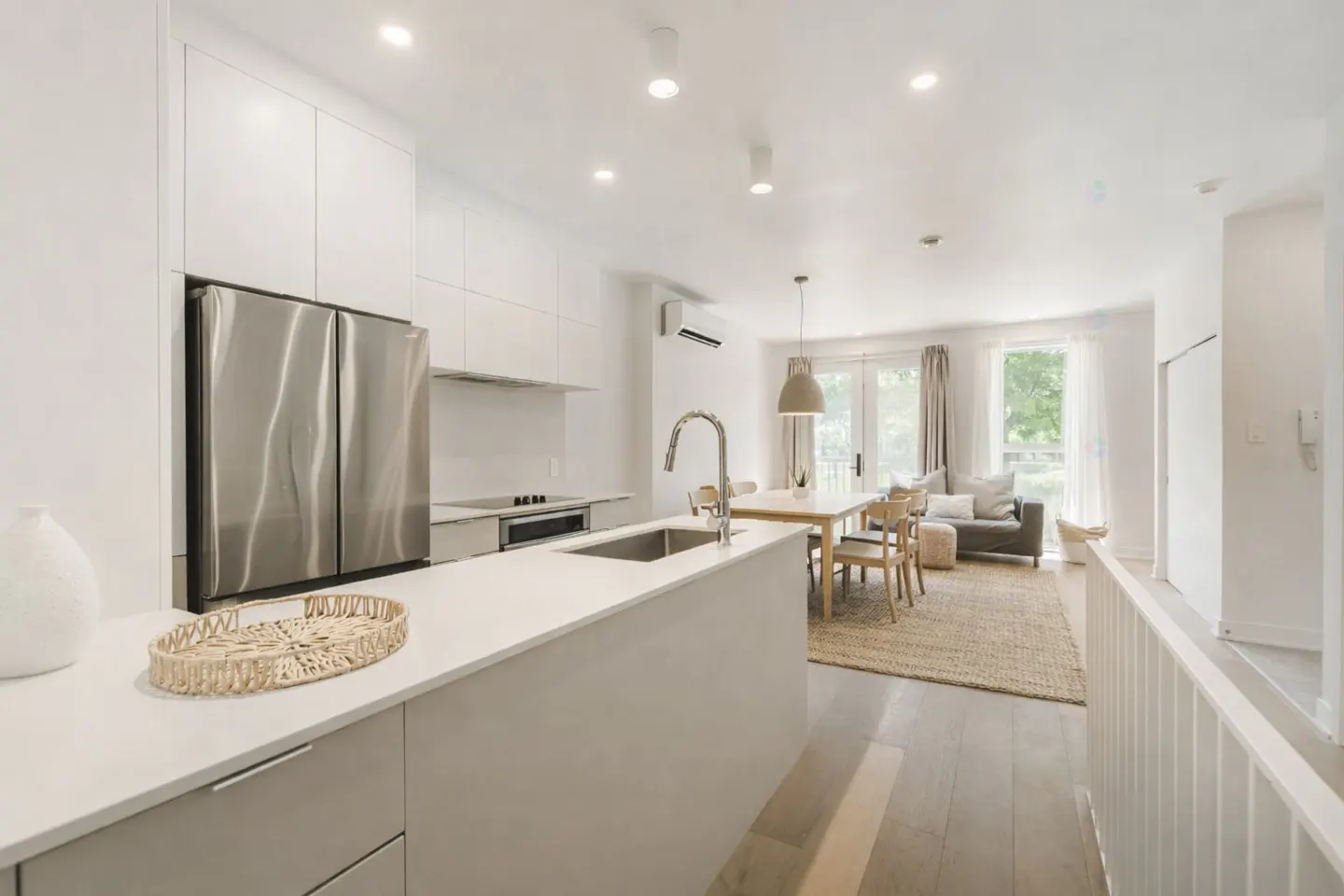 Bright, open-concept kitchen and living area. Stainless steel fridge, white cabinets, island with sink, dining table, and gray sofa.