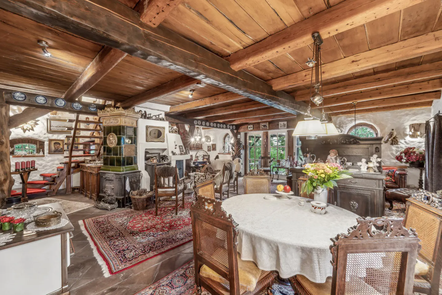A rustic dining room with a wooden beamed ceiling, an oval table with a white tablecloth, and ornate wooden chairs. A green tiled stove and a red rug add color.