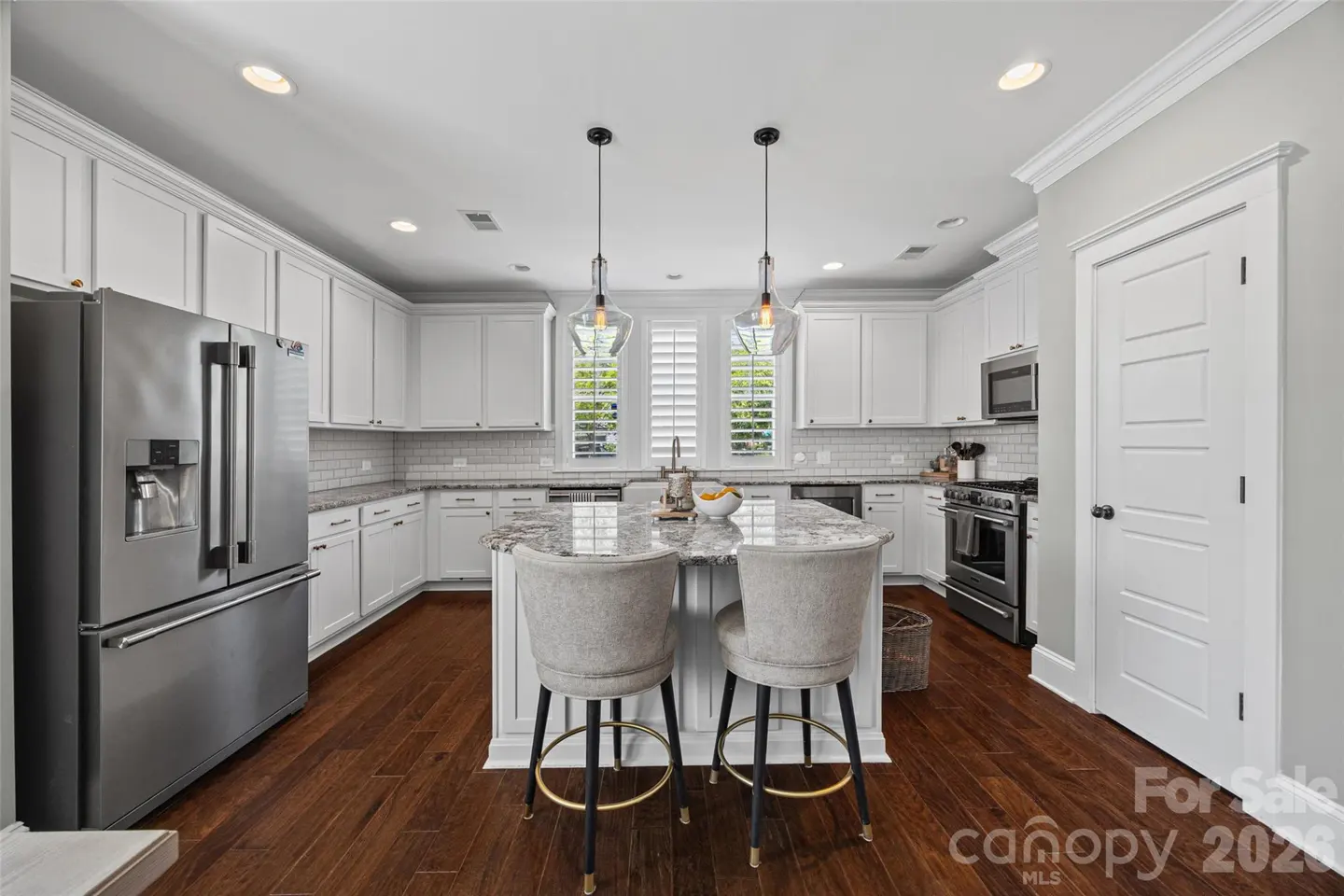 Modern kitchen with white cabinets, granite island, two grey bar stools, stainless steel appliances, and dark wood floors.