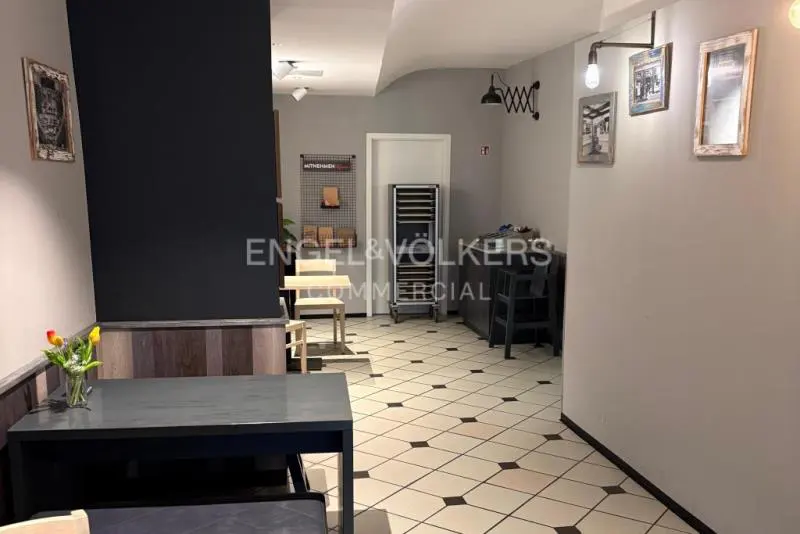 Interior view of a commercial space with black tables, chairs, and a black accent wall. The floor is tiled in a diamond pattern. Artwork hangs on the gray walls.