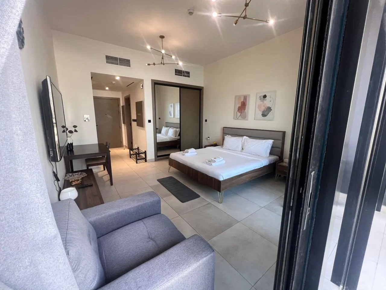 A modern studio apartment with a gray sofa, bed with white linens, and a dining table. The room has light beige walls and tile flooring.