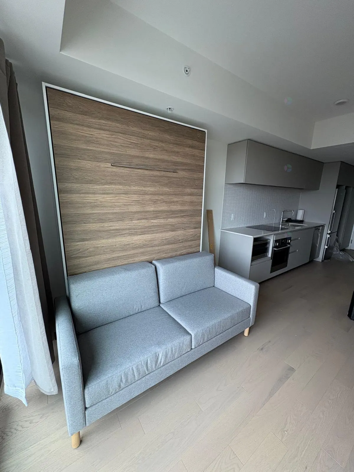 A gray sofa sits in front of a closed, light-wood Murphy bed in a modern apartment with a gray kitchen.