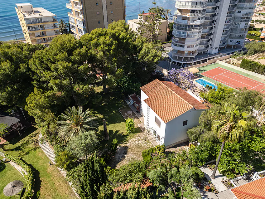 Aerial view of a white house with a red tile roof, surrounded by lush green trees, a tennis court, and the ocean in the background.