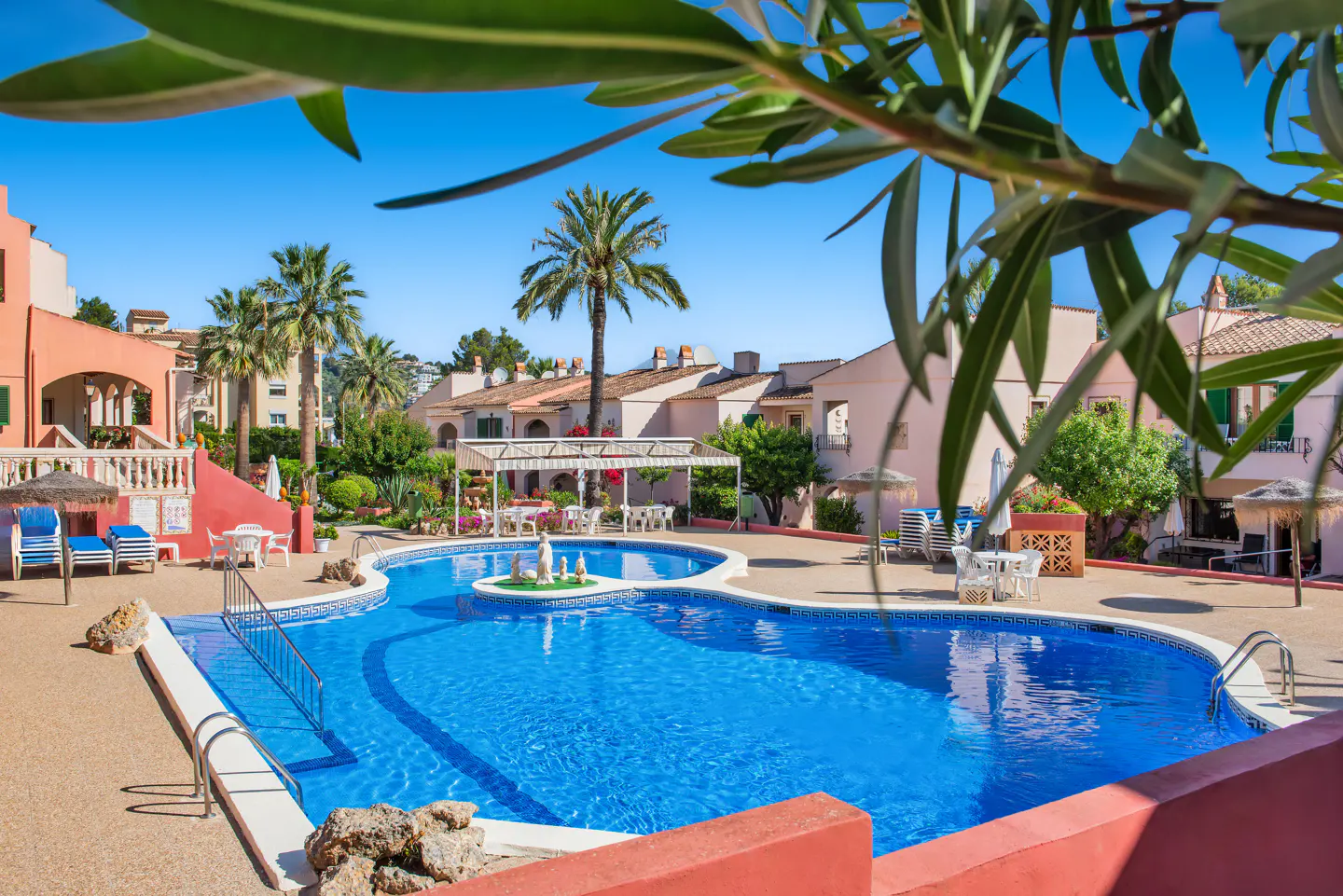 A bright blue swimming pool is surrounded by peach buildings and palm trees under a clear blue sky.