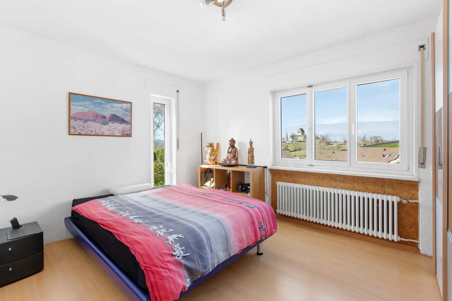 Bright bedroom with a blue-framed bed, red and grey patterned bedding, and a window view of a village. A wooden shelf holds Buddha statues.