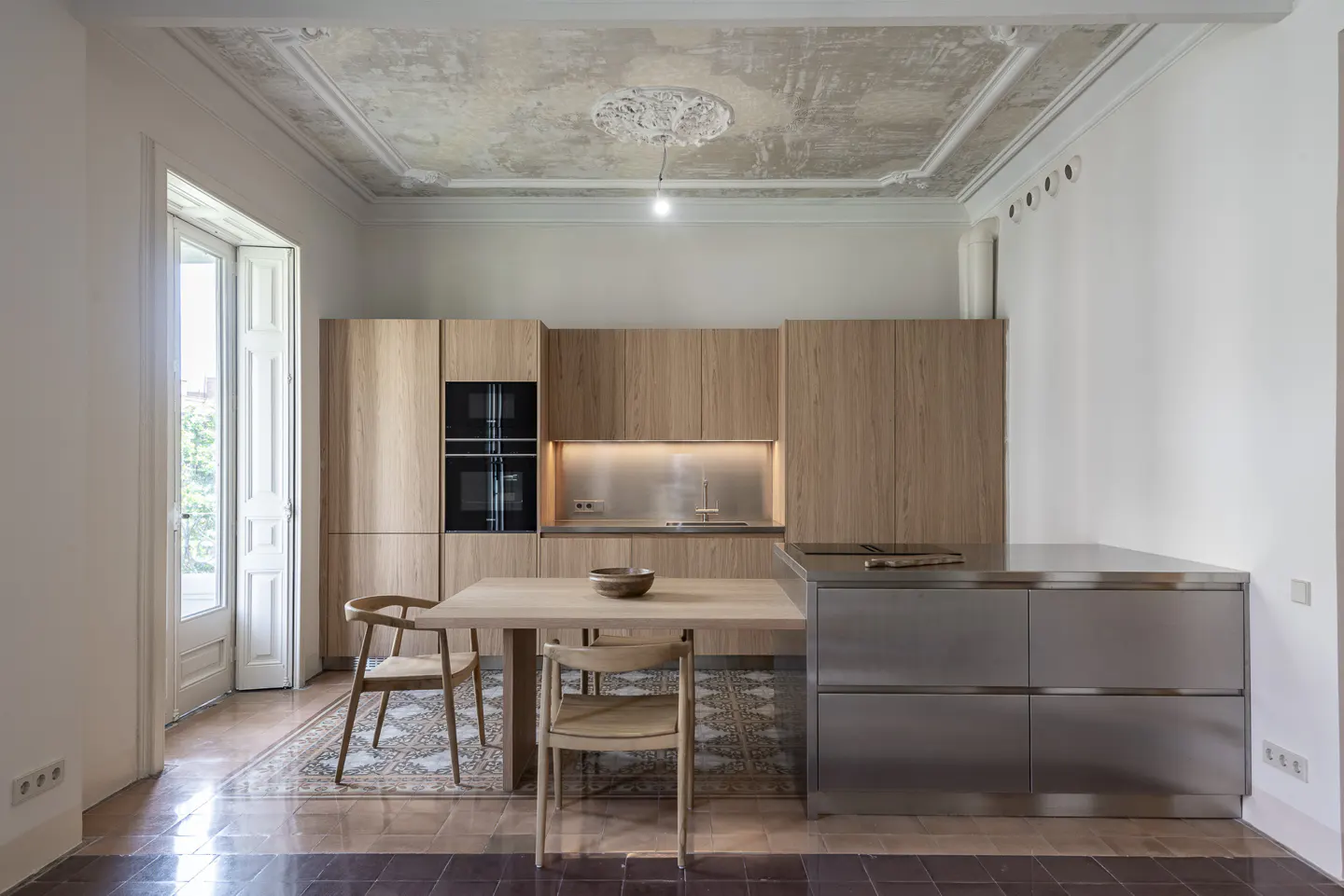 A modern kitchen with light wood cabinets, stainless steel island, and a wooden table with chairs. The floor is tiled, and a door leads to a balcony.