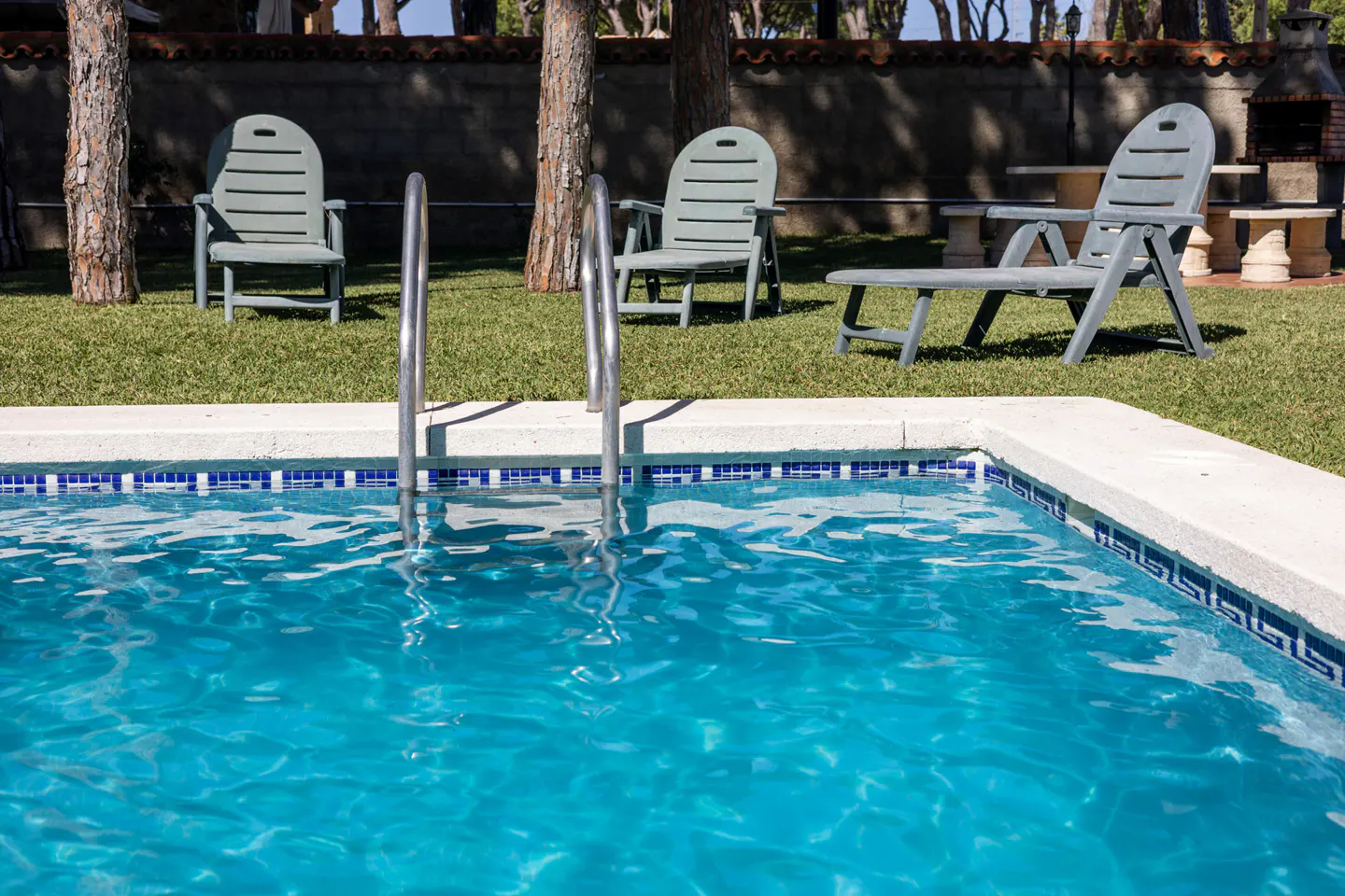 A backyard pool with blue water and a ladder, surrounded by green grass and three light green chairs.