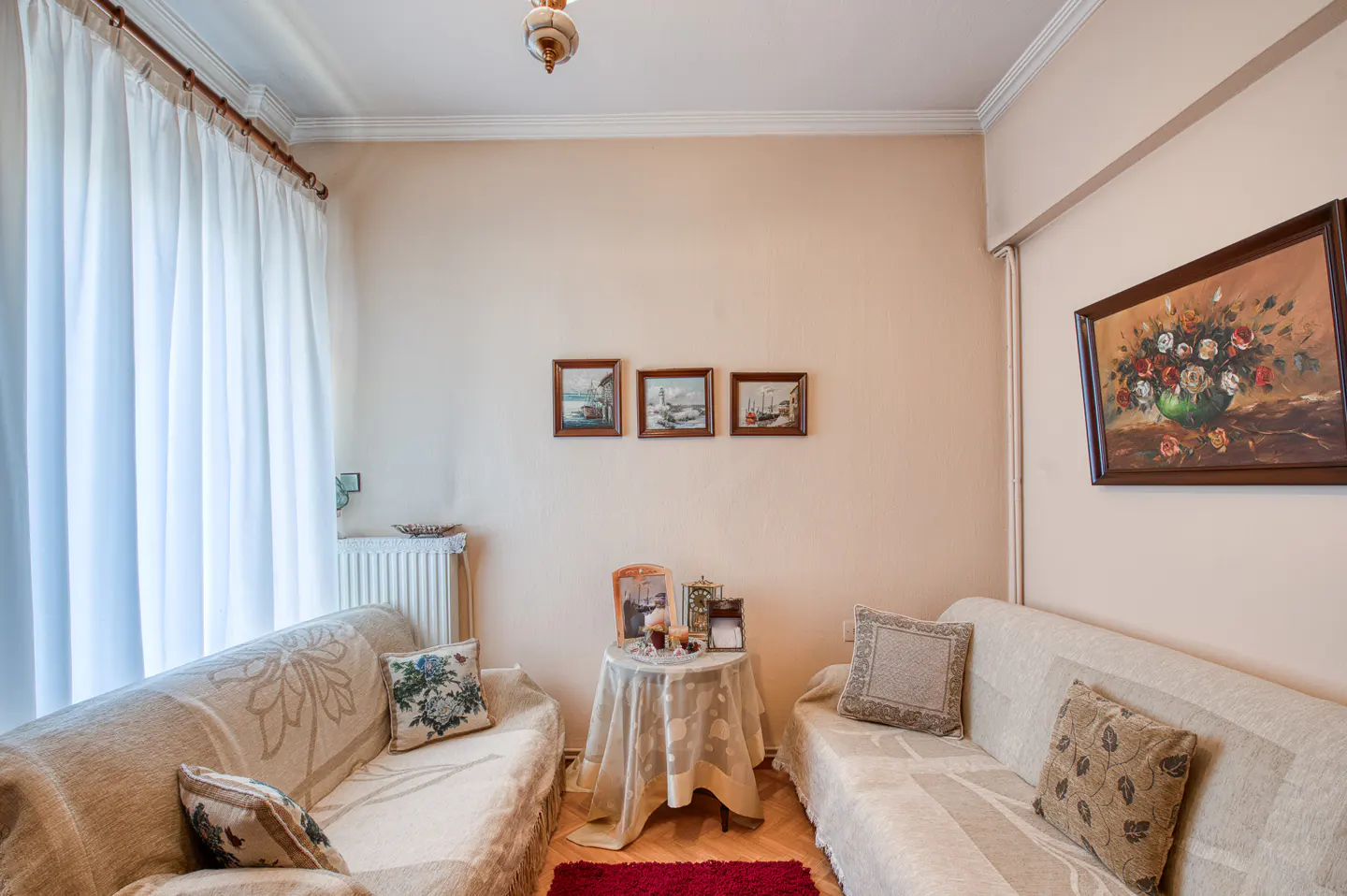 Living room with two sofas, a small table with objects, and framed art on the beige walls. White curtains hang by the window.