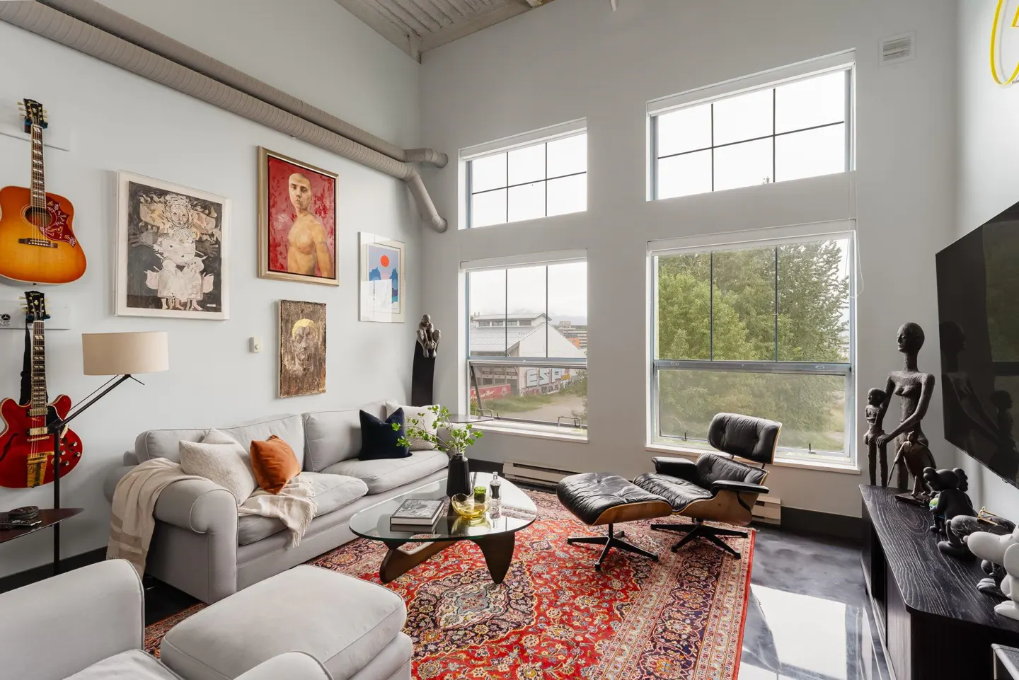 Bright living room with guitars on the wall, a gray sofa, and a black leather chair on a red patterned rug. Tall windows overlook a cityscape.