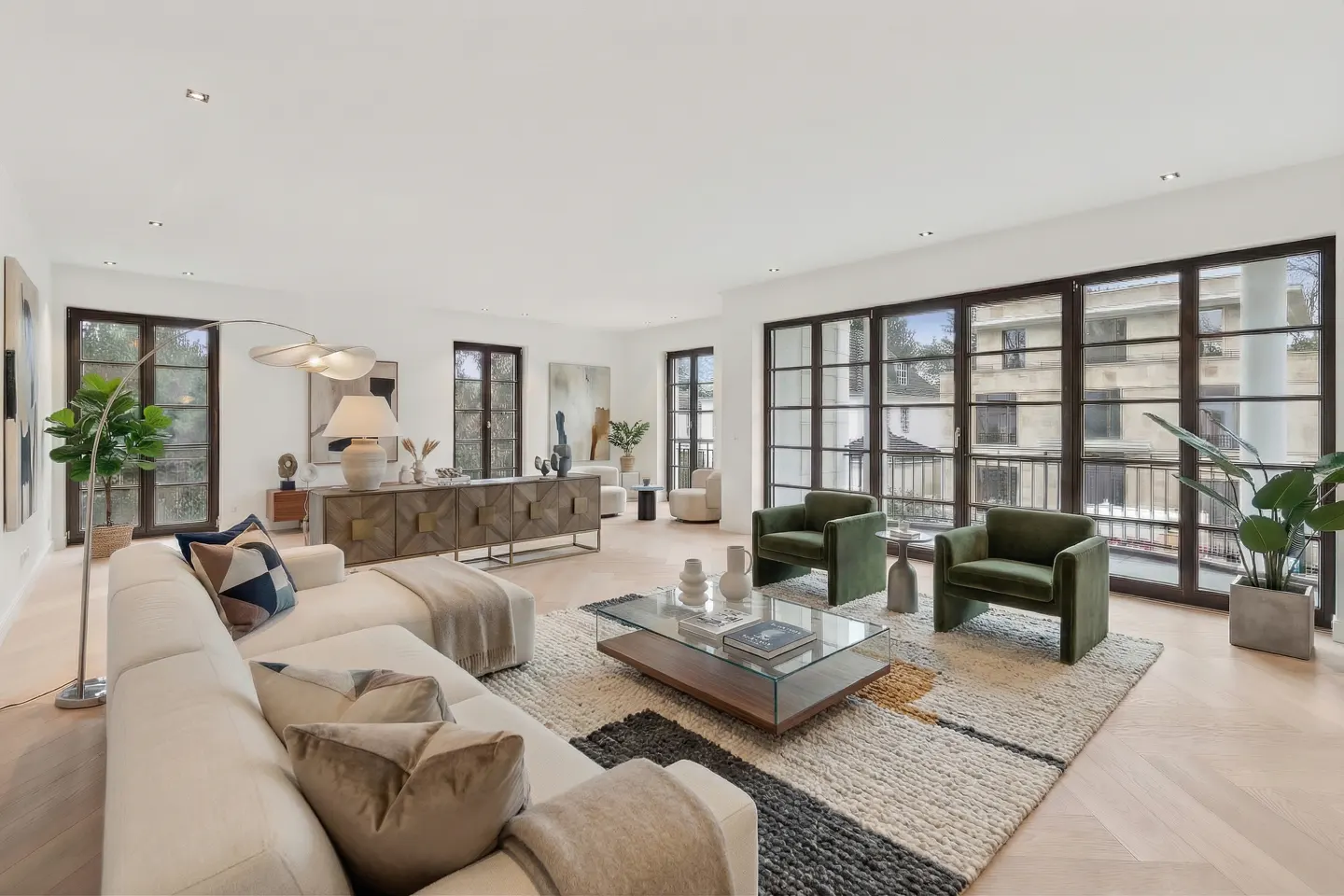 Modern living room with a white sectional, two green velvet armchairs, a glass coffee table, and large floor-to-ceiling windows.