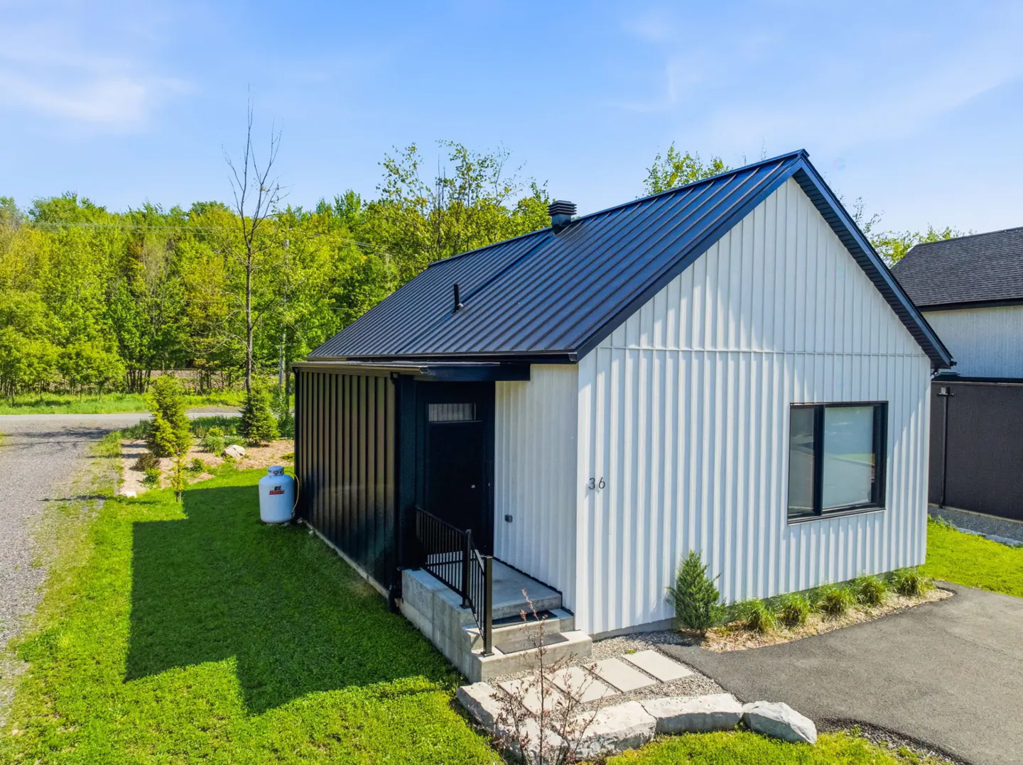 Modern home with white vertical siding and a black metal roof. A black door and small porch are visible. Green lawn and trees surround the house.