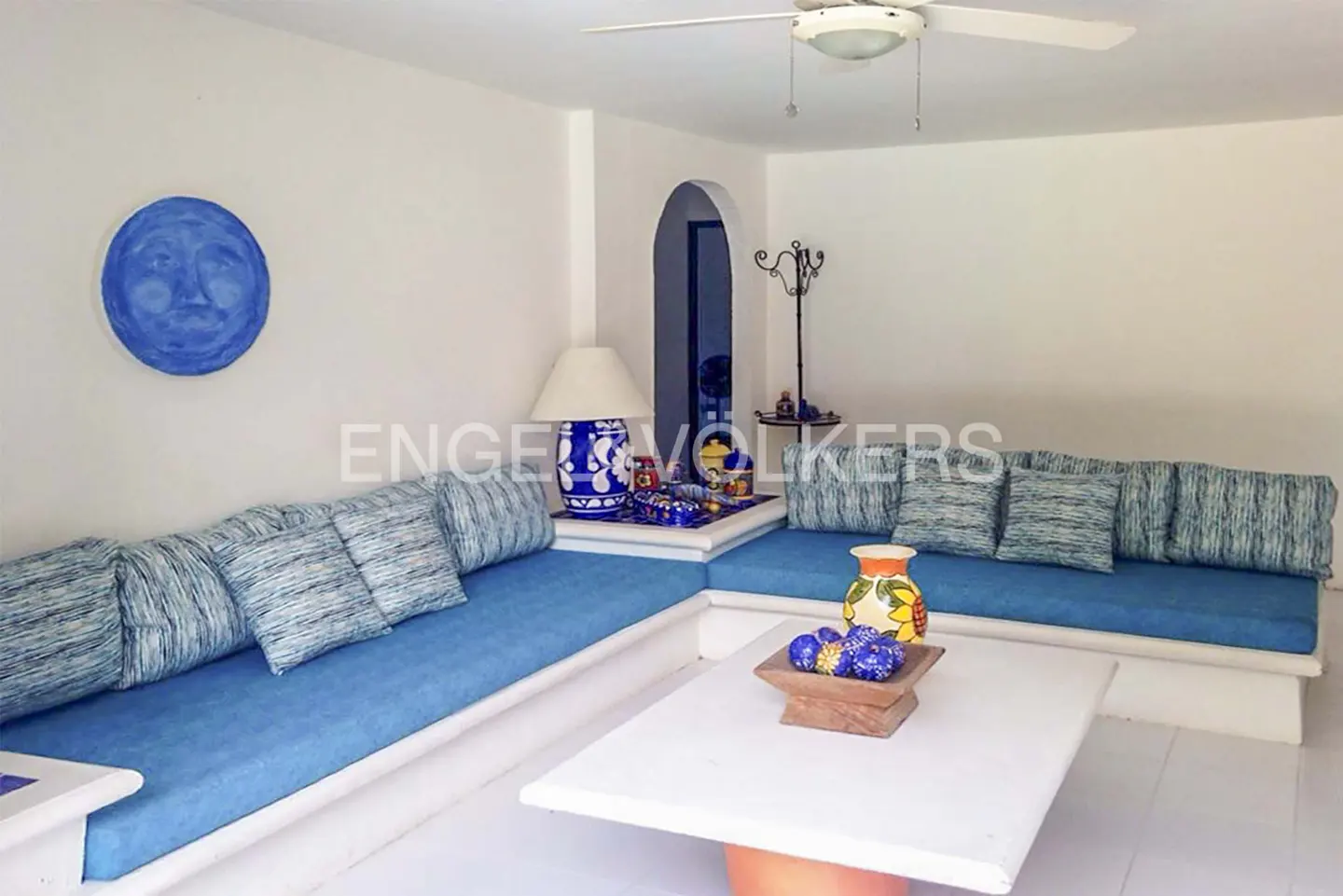 Bright living room with white walls, blue cushions on a built-in sofa, and a white coffee table. A blue moon art piece hangs on the wall.