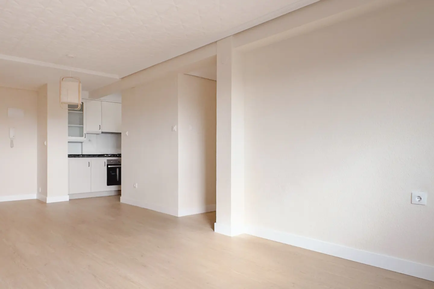 Bright, empty apartment with light wood floors and white walls. A kitchen with white cabinets is visible in the background.