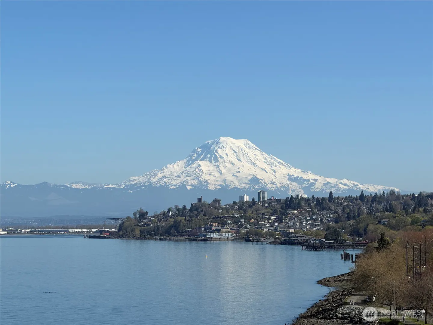 Scenic view of Mount Rainier, a snow-capped mountain, behind a city on the Puget Sound under a clear blue sky.