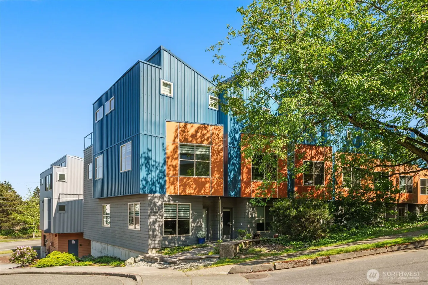Modern townhouses with blue and orange panels, gray siding, and white windows, set against a blue sky and green trees.