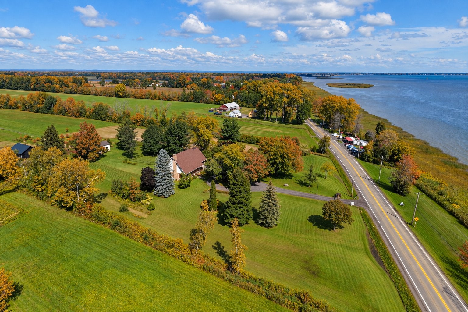 Aerial view of a house on a large green lawn next to a road, autumn trees, and a blue lake under a partly cloudy sky. [AI-generated text]