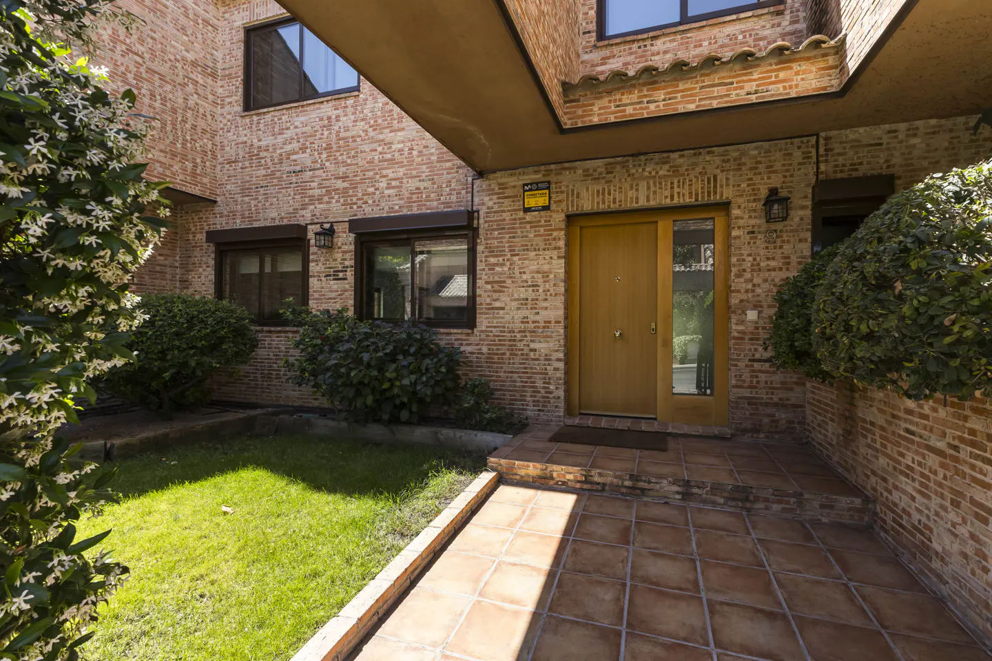 Brick house exterior with a wooden front door, brown window frames, and a tiled walkway leading to a green lawn.