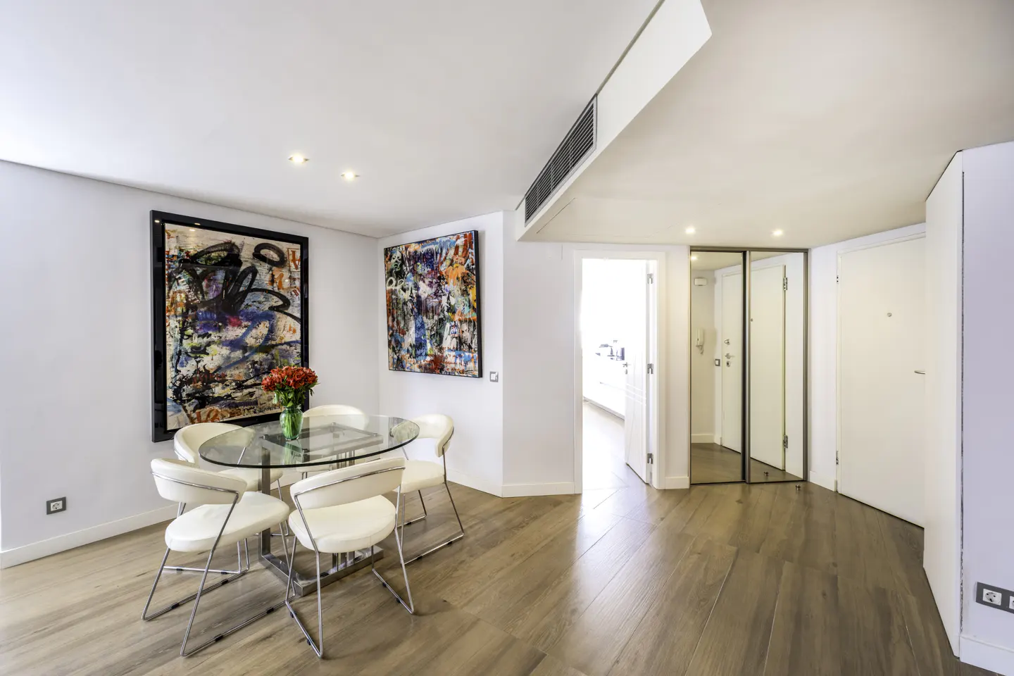 Bright dining area with wood floors, white walls, and abstract art. A glass table with white chairs sits near a vase of red roses.