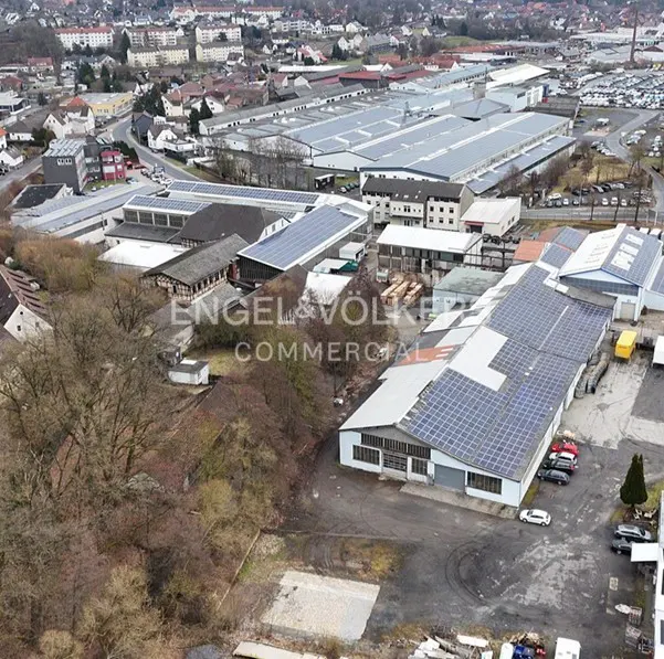 Aerial view of commercial buildings with solar panels on roofs, surrounded by trees and a cityscape in the background.