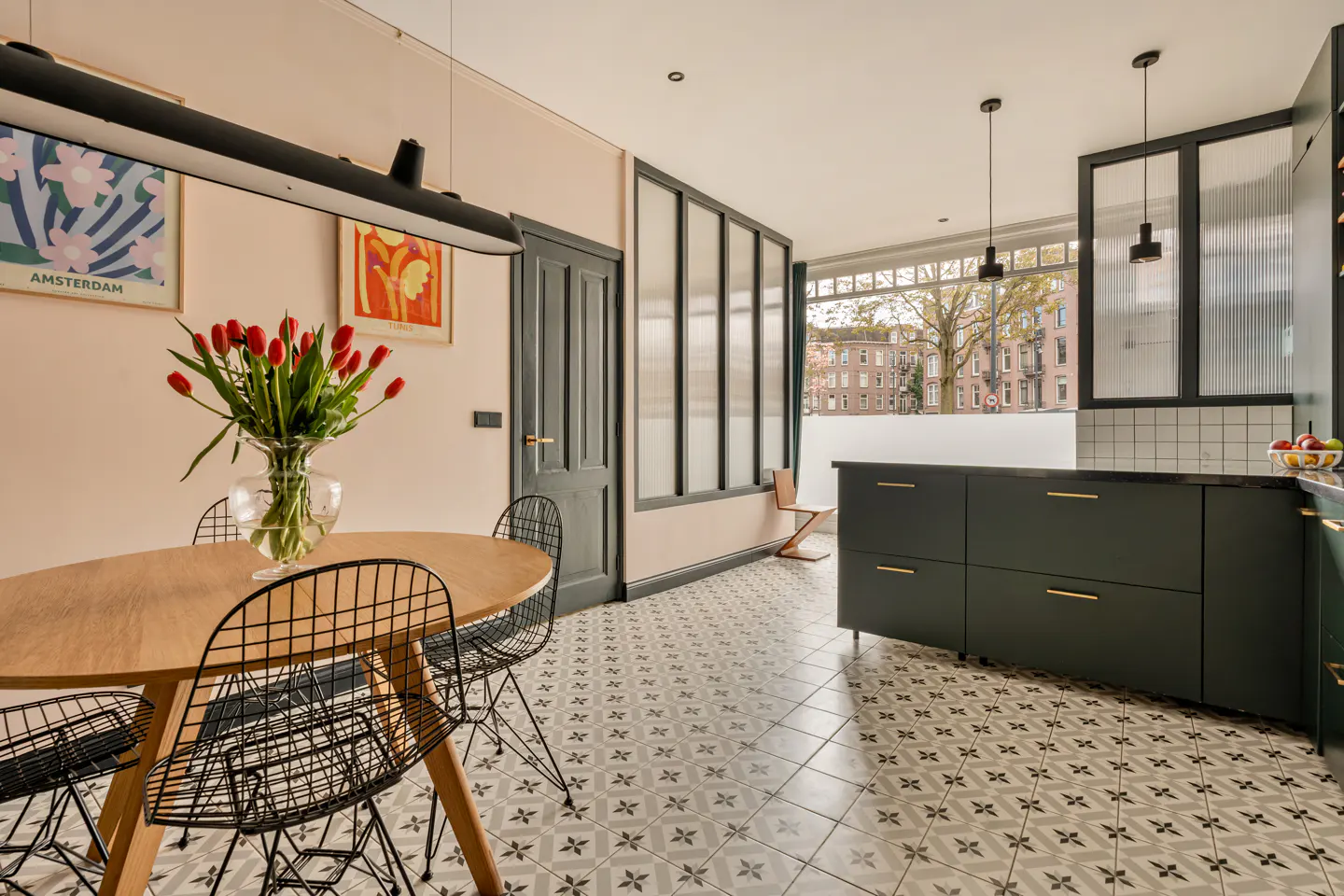 A modern kitchen with a round wooden table, black wire chairs, and a vase of red tulips. Dark green cabinets and patterned tile floor.