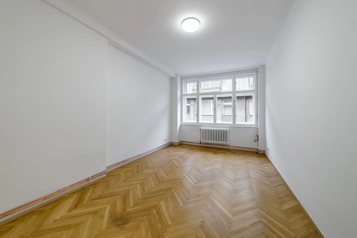 Bright, empty room with white walls, herringbone wood floor, and a large window with a radiator.