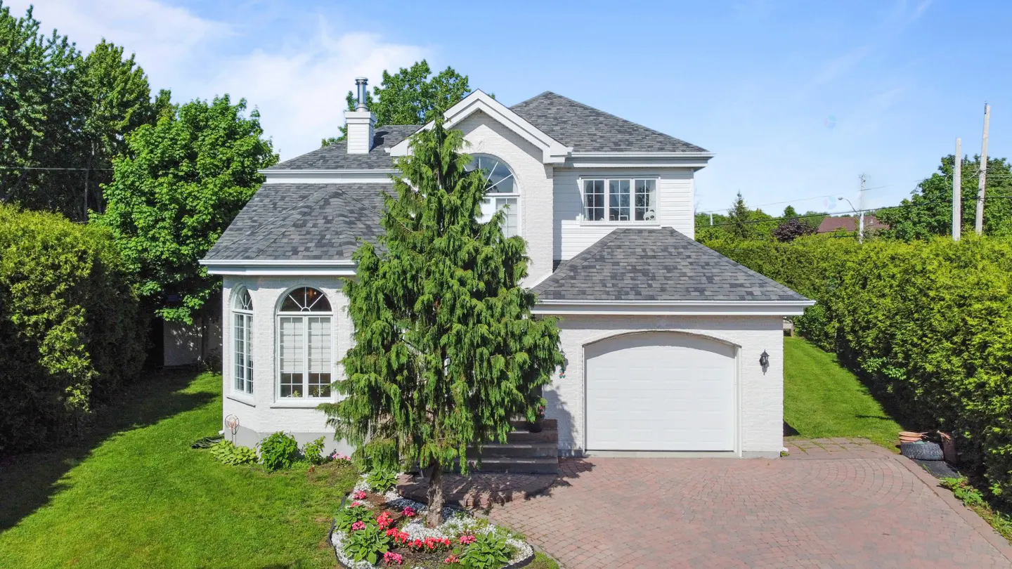 Two-story white house with a gray roof, green lawn, and brick driveway on a sunny day.