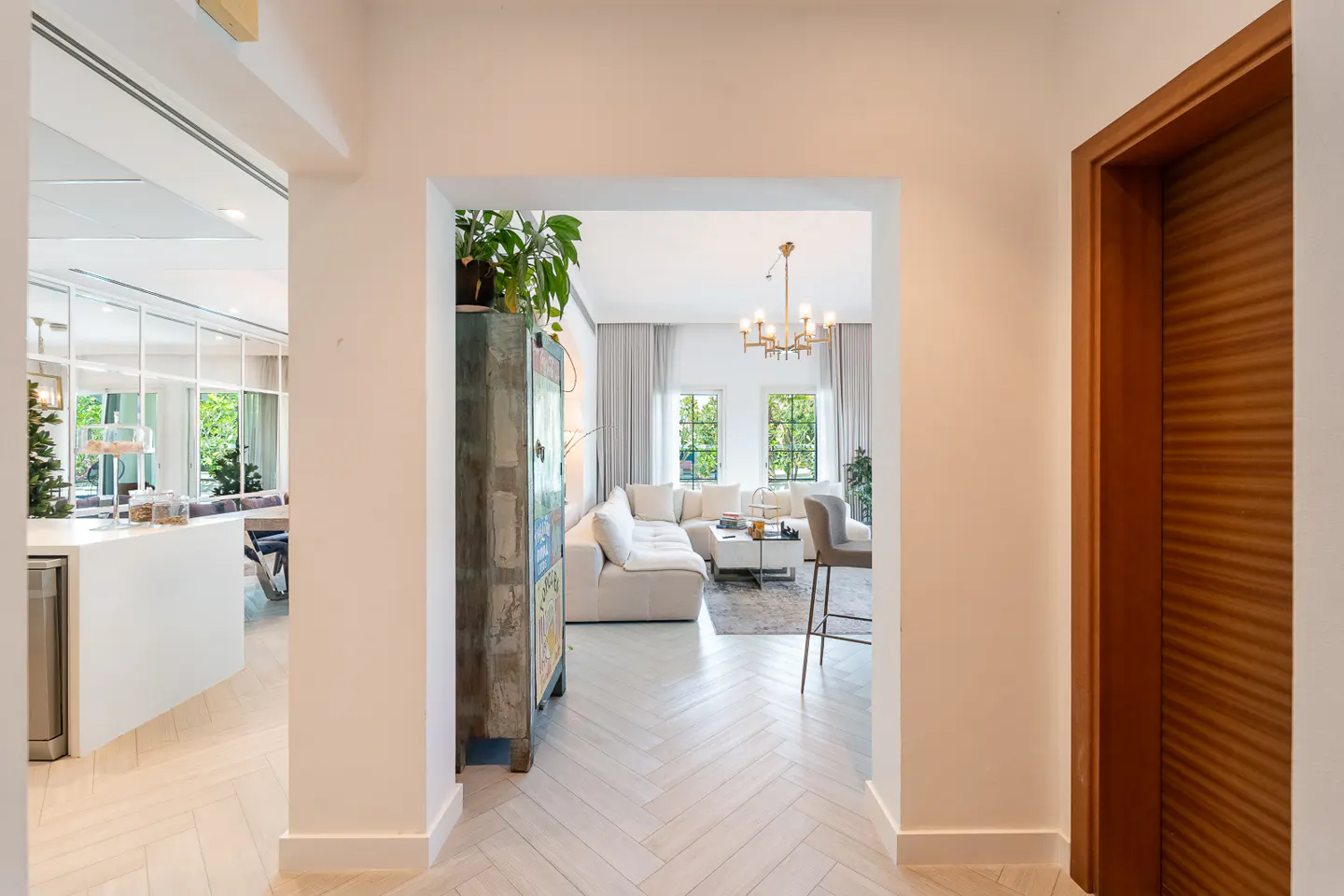 Hallway view into a bright living room with white walls, herringbone floors, and a brown wooden door on the right.