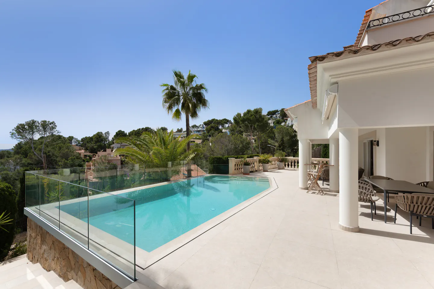 Luxury villa with a turquoise pool, glass railing, and outdoor dining area. Palm trees and lush greenery in the background under a clear blue sky.