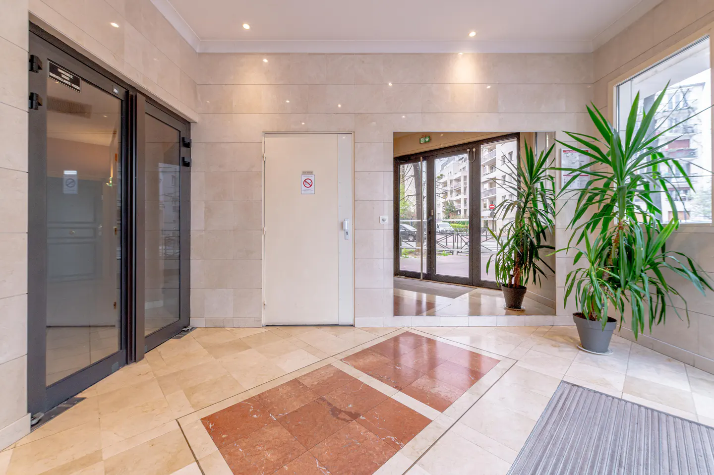 Modern apartment building lobby with beige tiled walls, glass doors, a central white door, and a large potted green plant.