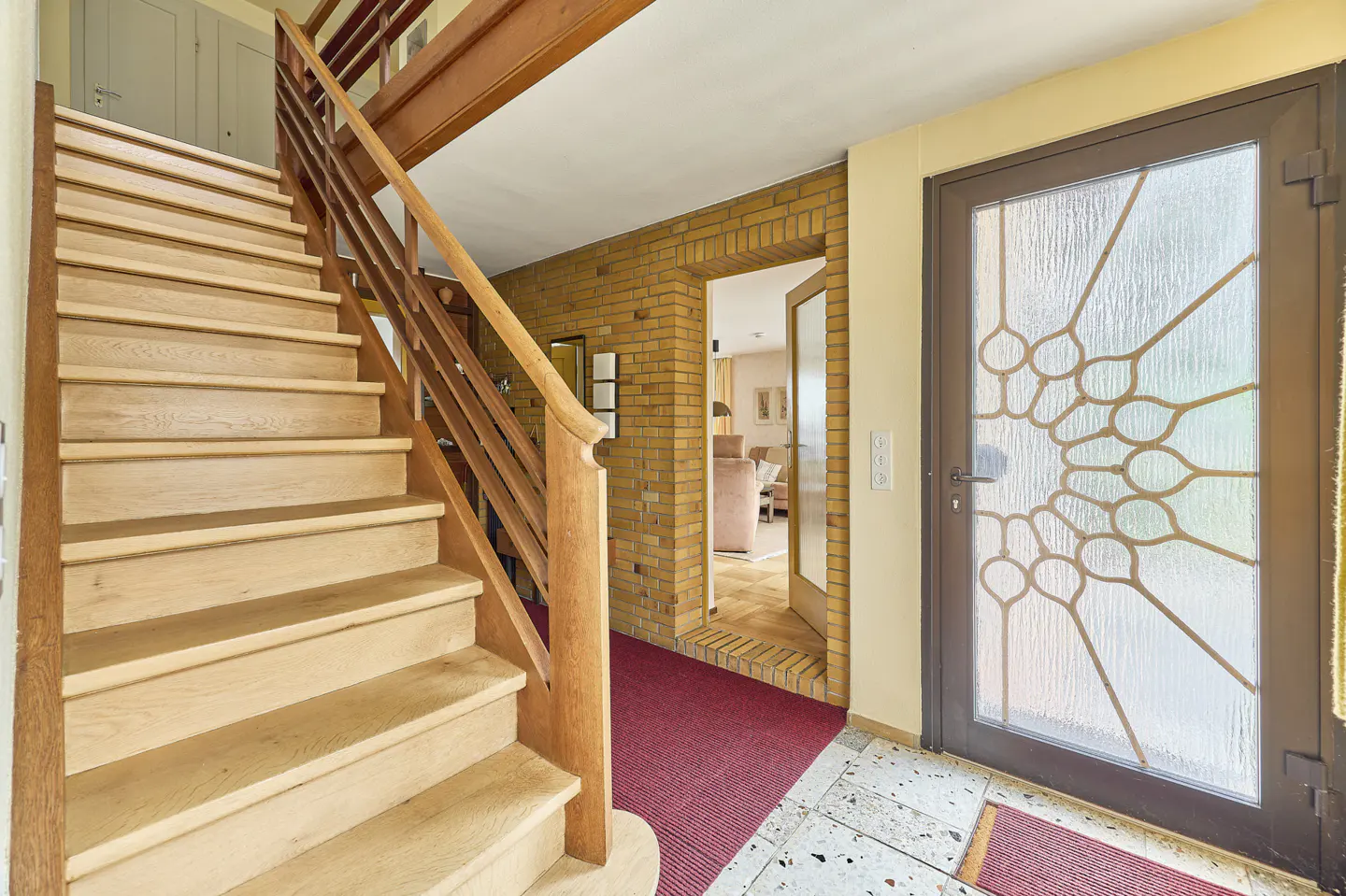 Entryway with light wood stairs, brick wall, and patterned glass door. A red rug leads to an open doorway revealing a glimpse of the living room.