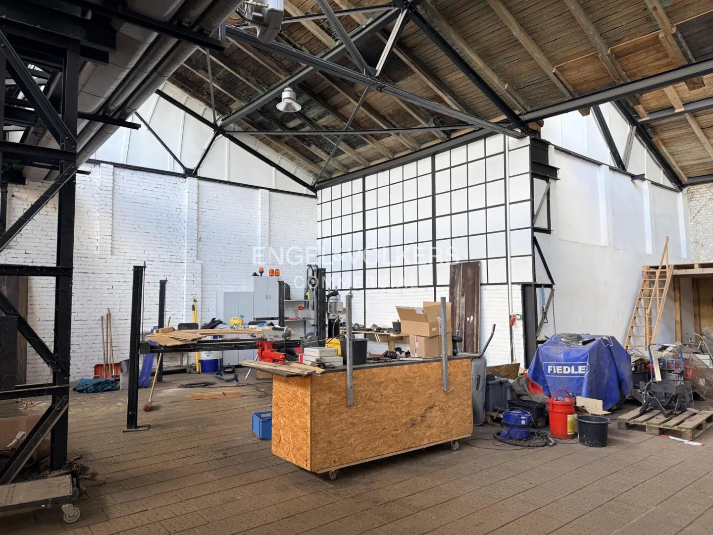 Interior of a large, unfinished warehouse space with exposed beams, white brick walls, and tools scattered around.
