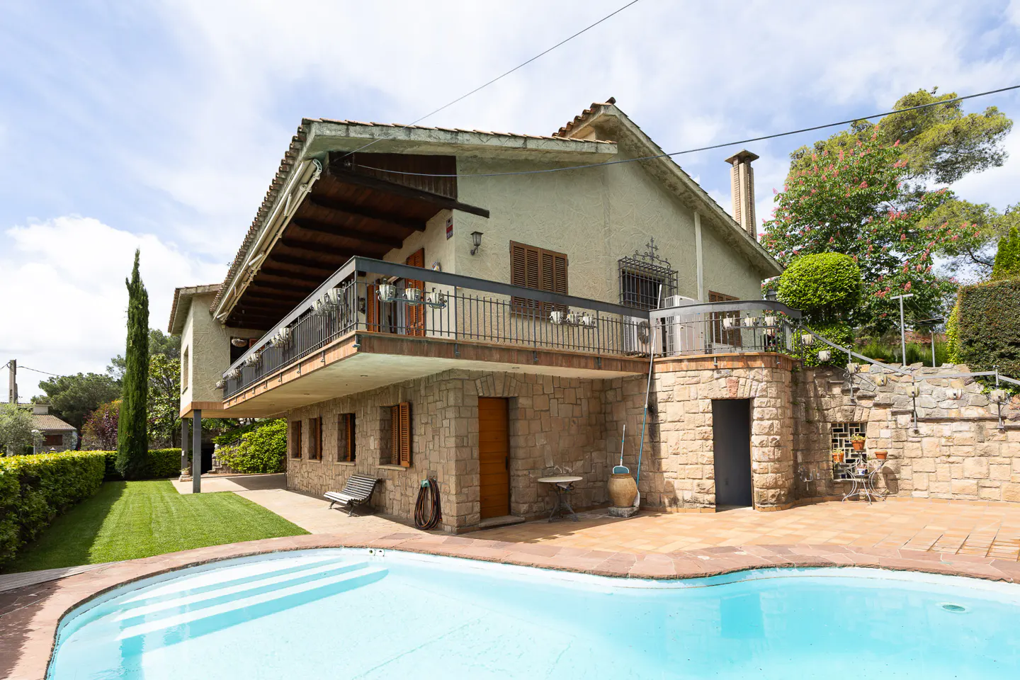 Exterior view of a two-story house with a pool, stone walls, and a balcony under a partly cloudy sky.