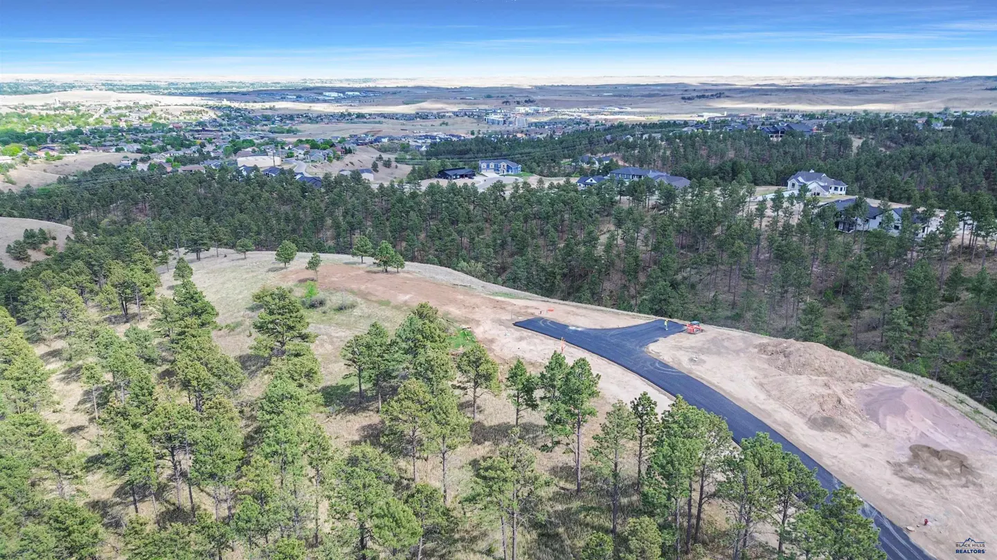 Aerial view of a paved T-intersection road under construction, surrounded by pine trees and a distant suburban landscape.