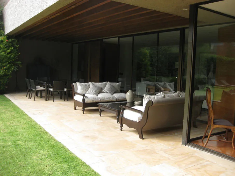 Outdoor patio with stone flooring, sofas, table, and dining set. Large glass doors reflect the green landscape.