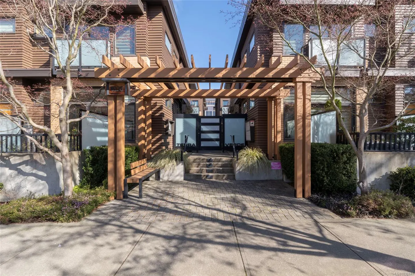 Exterior view of brown townhouses with a wooden pergola entrance, bench, and landscaped walkway.